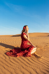 Fototapeta na wymiar Young beautiful woman in long red dress with red rose petals among the desert. Desert rose conception.