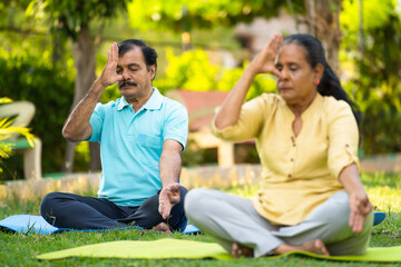 Indian senior couple doing pranayama or nostril breathing yoga during morning at park - concept of...
