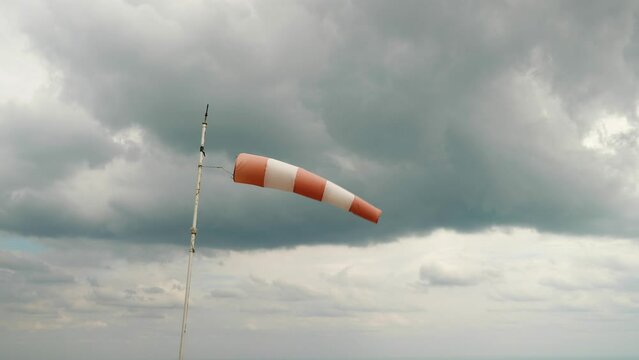 Against the cloudy sky, a white and red windsock dangles in the wind. Mounted on a pipe with peeling paint. It's a cloudy, windy day