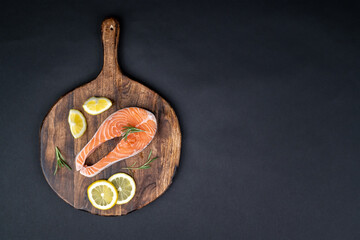 Fresh raw salmon slice with lemon and rosemary arranged on a wooden board on black background with copy space. Healthy seafood. Top view.