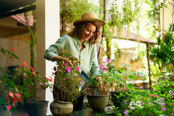 Young woman gardener in straw hat holding hand shovel taking care of potted plants. Junior caucasian female smiles standing in her little garden planting flowers in pots. Gardening and farming concept