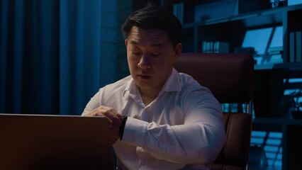 Adult Asian man Chinese worker Korean businessman working hard computer online laptop work project deadline late in home office tired exhausted looking at wristwatch check time evening overworked male