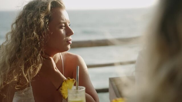 Blonde young woman in white dress in a sea view tropical cafe talks, smiles, wind blows her long hair. Happy young curly female chats with girls in seaside outdoor restaurant at sunset. Ocean view bar