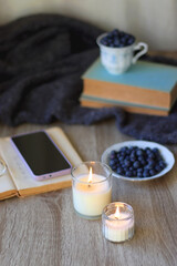 Cup of blueberries, set of books, phone, eyeglasses, lit candles and blanket on the table. Hygge at home. Selective focus.
