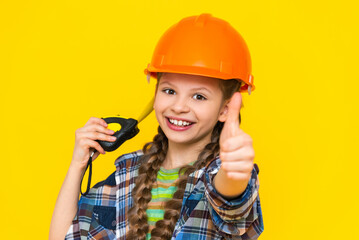 A teenage girl in a construction helmet shows a thumbs up sign and holds a tape measure. The child is an engineer. The concept of renovation in the children's room.