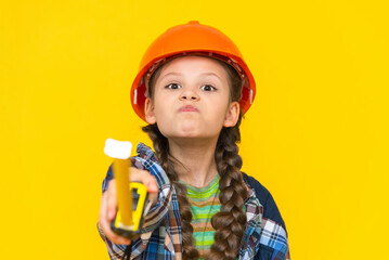 A disgruntled little girl builder in a construction helmet points forward with a tape measure. A harmful child. Renovation in the children's room. Yellow isolated background.
