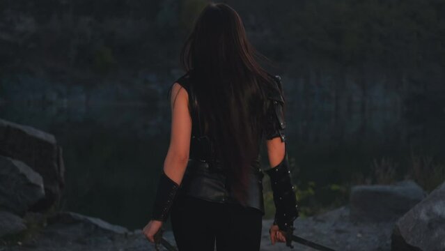 silhouette fantasy woman warrior assassin holds dagger in hands walks at night nature. Rear view, back armed girl. Black leather costume, hood. Ninja soldier with sword knife. 4k footage. river rocks.