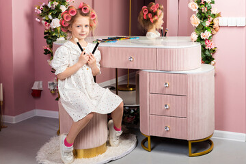 Little girl wearing hair rollers and white polka dot dress sits in the pastel pink dressing room by...