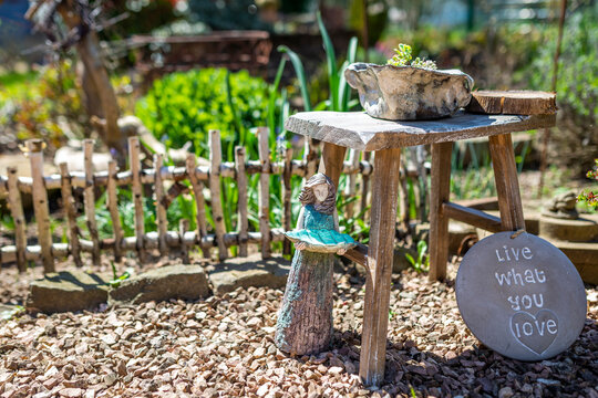 A small wooden figure holds a blue leaf and stands next to a wooden stool. there is a sign next to it that says: live what you love, in the garden