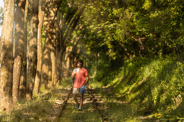 Obraz premium Backpacker man, walking along train tracks, observing Portugal's lush green forest in summertime. Wearing hat, pink t-shirt and short with sandals. Sunbeams.