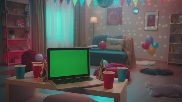 Laptop with green screen display in front of a room decorated for a house party. On a blurred background, multicolored lights flash and a disco ball sparkles. Advertising area, workspace mock up.