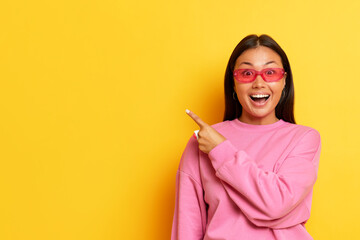 Cheerful dark-haired woman in pink sunglasses and pink sweatshirt stands on yellow studio backdrop,...