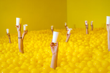 people's hands stick out of the pool with yellow balls, holding a white paper cup in their hand