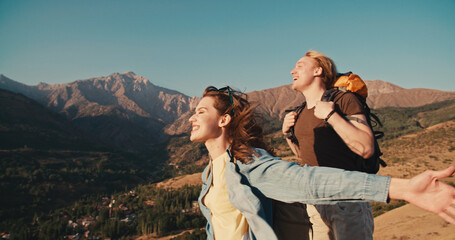 Man and woman hikers laughing with hands up, on a top of the mountain at sunset in mountains. Couple raising up hands on high rock in evening nature. Tourism, traveling and healthy lifestyle concept.