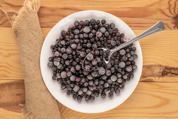 Frozen black currant with metal spoon and white plate on wooden table, macro, top view.