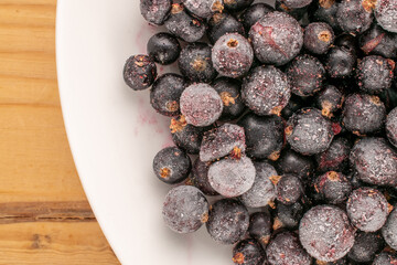 Frozen black currant with white plate on wooden table, macro, top view.