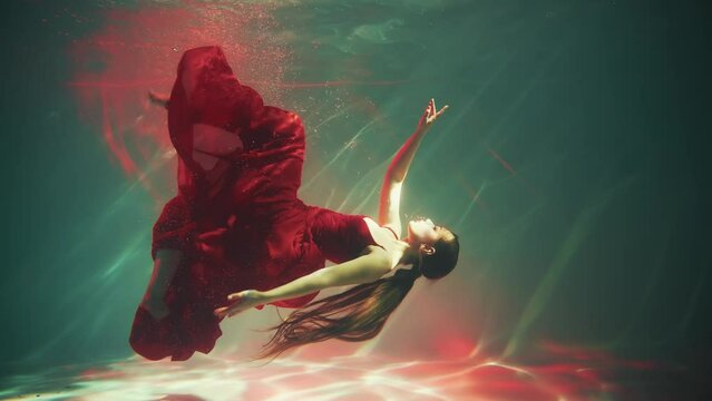 Fantasy woman mermaid swims floating in depth underwater world long red dress fabric tail in slow motion. Sexy girl princess of ocean sea dark blue water. art magic pool. wet lady nymph siren myth 4k
