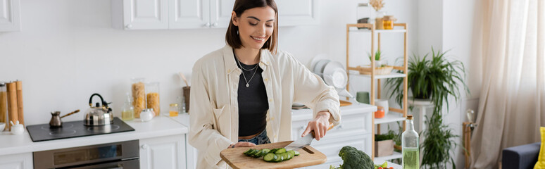 Cheerful woman holding sliced cucumber on chopping board in kitchen, banner.