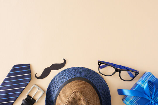 Father's Day atmosphere concept. Top view flat lay of trendy necktie hat glasses belt black mustache and gift box on light beige background with empty space for greeting text or advert