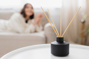 Bamboo sticks in aroma diffuser on coffee table near blurred woman in living room.