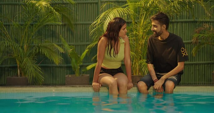 Happy young 2 Indian Asian guy couple sitting edge of swimming pool hot summer holiday relax talk together fun joy resort Boyfriend girl friend discuss plan look each other enjoy communication moment