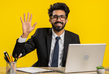 Indian business man working on laptop smiling friendly at camera, waving hands gesturing hello hi, greeting, goodbye, welcoming with hospitable expression at office workplace desk on yellow background © Andrii Iemelianenko