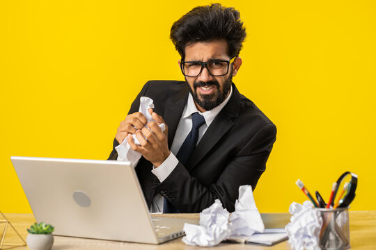 Angry furious indian man working at office throwing crumpled paper, having nervous breakdown at work, migaine, headache, stress management, mental distress problems, losing temper, reaction on failure