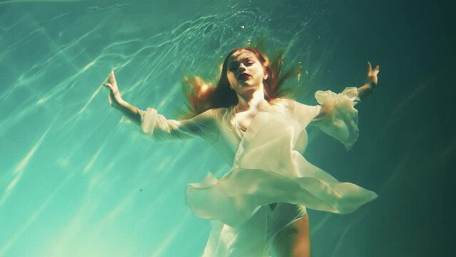 Fantasy woman drowns under dark blue water falls to bottom sea. Wet white silk long dress. Red-haired fairy girl goddess river nymph mermaid swims immersed. Creative underwater video shooting pool 4k