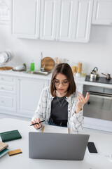 Obraz na płótnie Canvas Freelancer in eyeglasses using laptop near notebooks and credit card in kitchen.