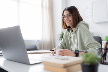 Cheerful freelancer in eyeglasses looking at laptop near coffee and books at home.