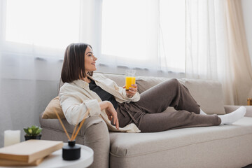 Side view of smiling brunette woman holding glass of orange juice while sitting on couch at home.