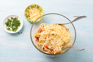 Glass bowl with summer pasta: spaghetti, tomatoes, red bell peppers, parsley, spices and grated cheese on a light blue background. cooking stage