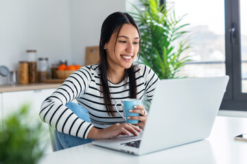 Smiling young woman doing video call with laptop while holding a cup of coffee in the kitchen at home. - obrazy, fototapety, plakaty