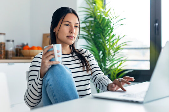 Smiling young woman doing video call with laptop while holding a cup of coffee in the kitchen at home.