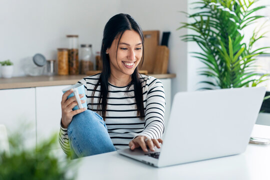 Smiling young woman doing video call with laptop while holding a cup of coffee in the kitchen at home.
