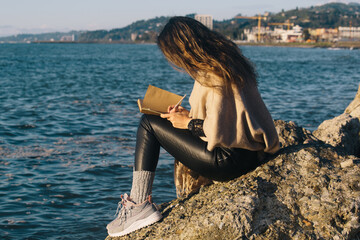 Girl sitting on stone at sea shore writing in notepad