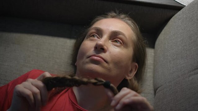 A woman in prostration lies on a sofa and twists a braided braid of hair in her hands
