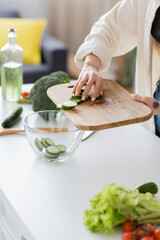 Cropped view of tattooed woman adding sliced cucumber in bowl in kitchen.