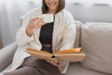 Cropped view of blurred woman holding coffee cup and reading book on couch at home.