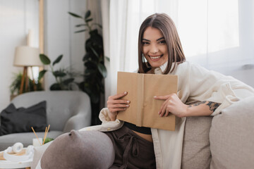 Smiling brunette woman holding book and looking at camera on couch at home.