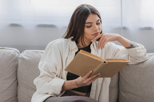 Tattooed young woman reading book on couch in living room.