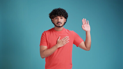 Young handsome latin spanish man with beard on blue studio background . Swearing with hand on chest and open palm, making a loyalty promise oath
