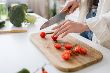 Cropped view of young woman cutting cherry tomatoes on chopping board in kitchen.
