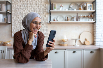 Close up photo. Smiling Muslim young woman in hijab standing in kitchen at home and using mobile...