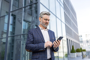 Fototapeta premium Portrait of a senior gray-haired male doctor standing in a suit outside the office and using a mobile phone, reading the news, typing a message, waiting for an appointment.