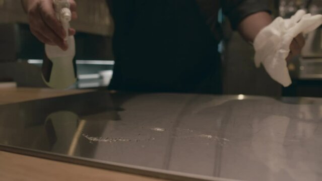 Close-up shot of hands of unrecognizable male chef dressed in uniform and apron cleaning in restaurant kitchen at end of shift, spraying counter with disinfectant liquid, and wiping with white cloth