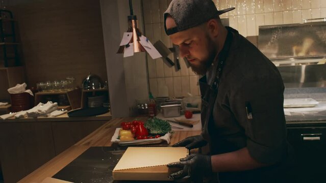 Medium shot of busy young Caucasian male chef in black uniform, apron and gloves, folding and closing takeaway box with freshly baked hot specialty pizza in dimly lit restaurant kitchen