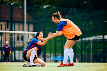 Happy female player helps her teammate to get up on soccer pitch.