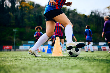 Unrecognizable woman with soccer ball running around cones during practice at stadium.