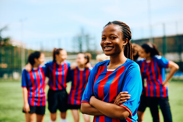 Happy black soccer player standing with her arms crossed on playing field and looking at camera.